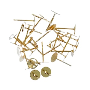 May include: A pile of gold-toned earring findings with a round base and a straight post. The findings are scattered on a white background.