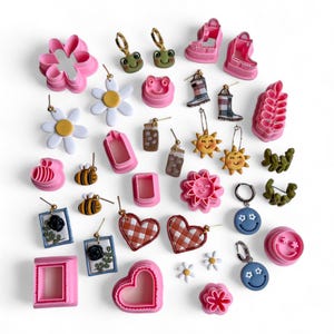 Peut inclure: Une collection de boucles d'oreilles en pâte polymère rose et verte de différentes formes, notamment des fleurs, des cœurs, des soleils, des abeilles, des grenouilles et des visages souriants. Les boucles d'oreilles sont présentées sur un fond blanc.