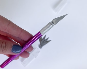 May include: A purple and silver craft knife with a retractable blade. The knife is held in a hand.