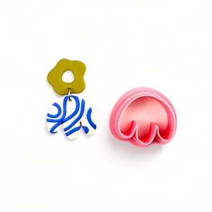 May include: A pair of earrings with a green, white, and blue abstract design. The earrings are made of polymer clay and are hanging from a silver hook. The earrings are next to a pink plastic cookie cutter in the shape of a flower.