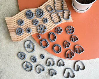 May include: A set of grey plastic cookie cutters in various shapes, including flowers, leaves, and abstract shapes. The cutters are arranged on a wooden surface with a brown background.