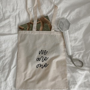 K&ouml;nnte beinhalten: Eine beige Tragetasche mit schwarzem Text, der "We are one" lautet. Die Tasche liegt auf einem wei&szlig;en Bettlaken. Ein wei&szlig;er Lichtschl&auml;ger mit dem aufgedruckten Wort "SEHUN" liegt rechts neben der Tasche auf dem Bettlaken.