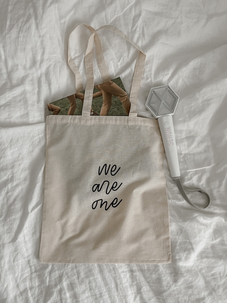 K&ouml;nnte beinhalten: Eine beige Tragetasche mit schwarzem Text, der "We are one" lautet. Die Tasche liegt auf einem wei&szlig;en Bettlaken. Ein wei&szlig;er Lichtschl&auml;ger mit dem aufgedruckten Wort "SEHUN" liegt rechts neben der Tasche auf dem Bettlaken.