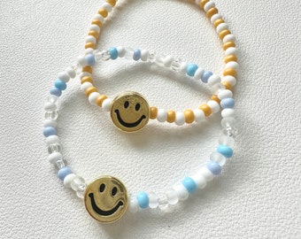May include: Two beaded bracelets with smiley face charms. One bracelet is white and blue with clear beads, the other is yellow and white.