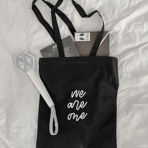 May include: Black tote bag with white embroidered text "We are one". The bag is filled with a notebook and a small black box with the word "EXO" on it. A white light stick with a K-pop logo is on the left side of the bag.