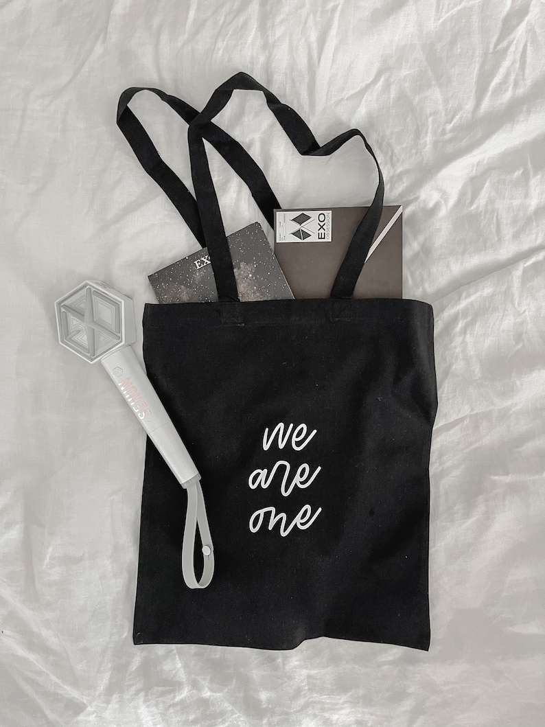 K&ouml;nnte beinhalten: Schwarze Tragetasche mit wei&szlig; gesticktem Text "We are one". Die Tasche ist gef&uuml;llt mit einem Notizbuch und einer kleinen schwarzen Box mit dem Wort "EXO" darauf. Ein wei&szlig;er Lichtschl&auml;ger mit einem K-Pop-Logo befindet sich auf der linken Seite der Tasche.