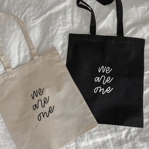 K&ouml;nnte beinhalten: Zwei Einkaufstaschen mit dem Aufdruck "We are one". Eine Tasche ist beige und die andere schwarz.