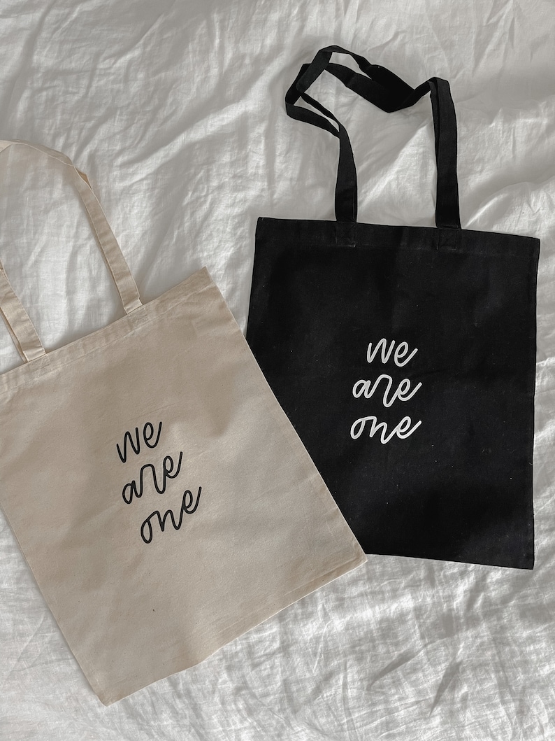 K&ouml;nnte beinhalten: Zwei Einkaufstaschen mit dem Aufdruck "We are one". Eine Tasche ist beige und die andere schwarz.