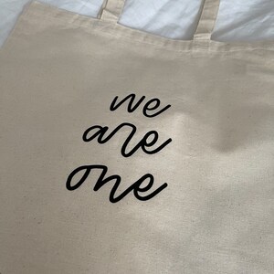 K&ouml;nnte beinhalten: Eine beige Canvas-Tragetasche mit schwarzen Henkeln und dem Text "we are one" in schwarzer Kursivschrift.