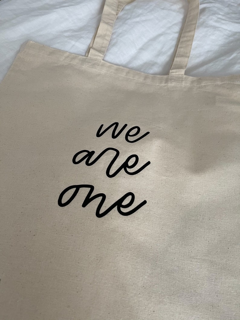 K&ouml;nnte beinhalten: Eine beige Canvas-Tragetasche mit schwarzen Henkeln und dem Text "we are one" in schwarzer Kursivschrift.