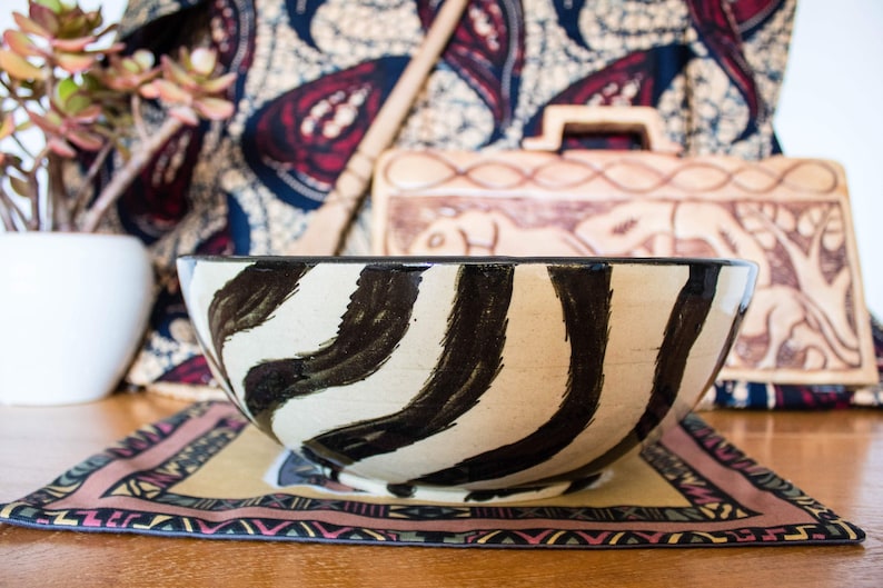 May include: A large ceramic bowl with a white background and black zebra-like stripes. The bowl is sitting on a colorful patterned placemat.