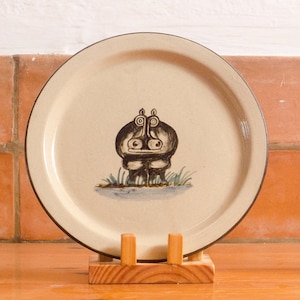 May include: A ceramic plate with a brown rim and a beige centre. The plate features a hand-painted illustration of a hippopotamus in black and brown.