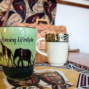 Może przedstawiać: Dwa ceramiczne kubki na stole z wzorzystą serwetką. Kubek po lewej stronie jest zielony z czarnymi sylwetkami żyrafy i słonia oraz napisem "Running Lifestyle". Kubek po prawej stronie jest biały z brązowo-czarną wzorzystą wstążką wokół górnej części.