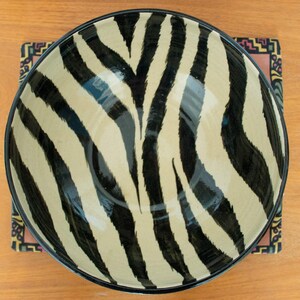 May include: A large ceramic bowl with a black and white zebra print design. The bowl is sitting on a colorful patterned placemat.