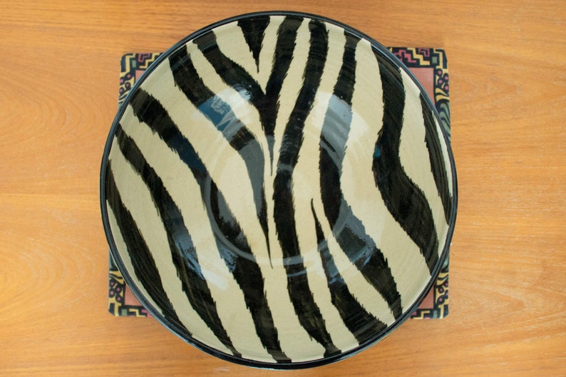 May include: A large ceramic bowl with a black and white zebra print design. The bowl is sitting on a colorful patterned placemat.