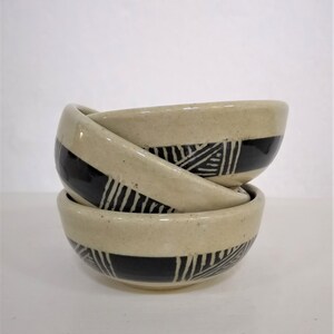 May include: Three ceramic bowls stacked on top of each other. The bowls are cream colored with black geometric designs.