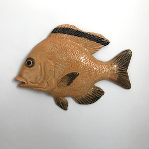 Puede incluir: Una decoración de pared de cerámica en forma de pez con un esmalte marrón claro y detalles negros. El pez tiene un diseño detallado con escamas y aletas.