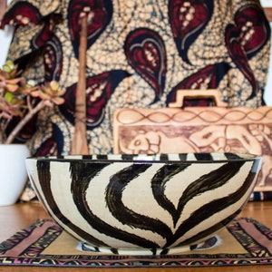 May include: A large ceramic bowl with a zebra print design. The bowl is sitting on a table with a colorful patterned placemat. The bowl is in focus, while the background is blurred.