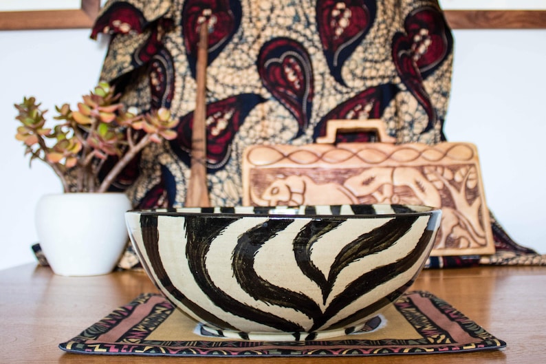 May include: A large ceramic bowl with a zebra print design. The bowl is sitting on a table with a colorful patterned placemat. The bowl is in focus, while the background is blurred.