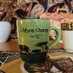 May include: A green ceramic mug with a black silhouette of animals and birds. The mug has the text "Chifuno Changa" written on it.