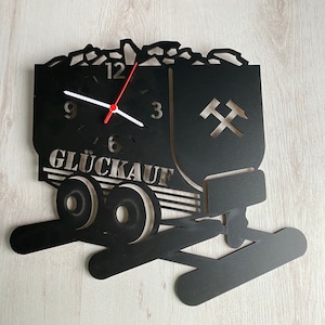 Könnte beinhalten: Schwarze Wanduhr aus Metall in Form eines Grubenwagens mit einem roten und weißen Zifferblatt. Das Zifferblatt hat die Zahlen 12, 3, 6 und 9. Der Wagen ist mit Kohle gefüllt und hat ein Pickel-Symbol an der Seite. Die Uhr zeigt "Glückauf", einen deutschen Bergmannsgruß.
