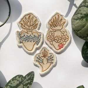 Op de afbeelding: Drie geborduurde patches in de vorm van een hart. De patches zijn beige met gouden, blauwe en rode steken. Eén hart heeft een doornenkroon, een ander bloemen en de derde een kroon en pijlen. Decoratieve textielkunst.