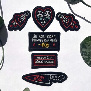 Op de afbeelding: Een verzameling zwarte geborduurde patches met rode en witte accenten. Ontwerpen omvatten een hart met een mes, een huilend hart, een mes en tekst die luidt "Se Son Rose, Punger Anno" en "Hello I'm dead inside."