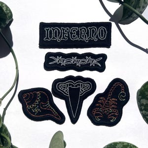 Può includere: Una collezione di toppe nere ricamate con cuciture bianche. Le toppe includono la parola "INFERNO", filo spinato, un serpente, una conchiglia e uno scorpione. Le toppe sono su una superficie bianca.