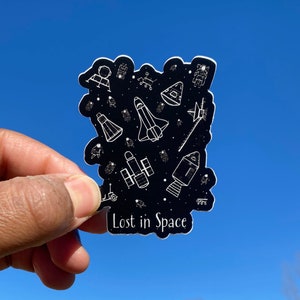 Op de afbeelding: Een zwart-witte sticker met een ruimtethema. Het bevat verschillende ruimte-illustraties, waaronder raketten, astronauten en planeten. De sticker heeft de tekst "Lost in Space" onderaan.