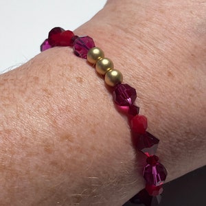 May include: A beaded bracelet featuring faceted red and magenta crystals interspersed with three gold-colored beads. The bracelet is worn on a wrist, showcasing the jewelry's design and color.
