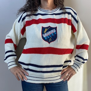 May include: Cream-colored knit sweater with red and navy blue stripes. The sweater features a shield-shaped patch with the words "Rhythm Blues" and a musical instrument design. The sleeves and neckline have navy blue stripes.