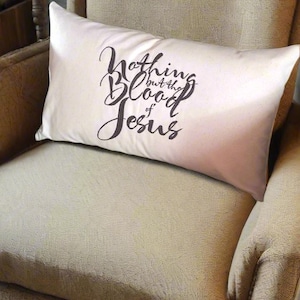 May include: A white throw pillow with the text "Nothing but the Blood of Jesus" in black cursive lettering. The pillow is sitting on a beige armchair.