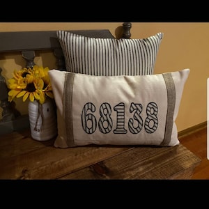 May include: A white pillow with a burlap trim and the number "68138" embroidered in black and white stripes. The pillow is sitting on a wooden surface.