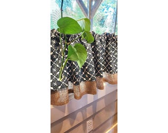 May include: A black and white patterned valance with a gold-colored bottom trim. The valance is hanging on a window sill, with a green plant growing through it. The window is visible in the background.