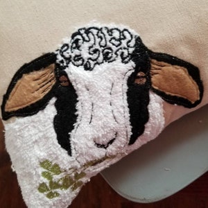 May include: A white and black embroidered sheep with brown ears, holding a green sprig in its mouth. The sheep is on a white background.