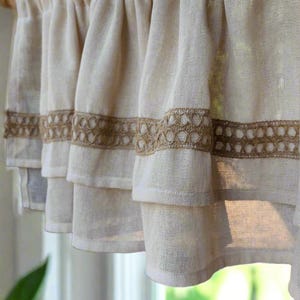 May include: A white tiered valance with a brown crocheted trim. The valance is made of a lightweight fabric and has a ruffled edge.