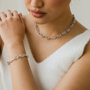 Könnte beinhalten: Eine silberne Kettenkette und ein Armband mit einem Ankerketten-Design. Die Kette wird von einer Person mit einem weißen, flauschigen Pullover getragen.