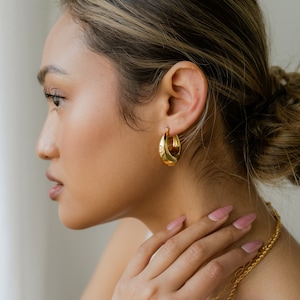 May include: A woman wearing gold hoop earrings with a textured design. She is wearing a white top and has her hair pulled back in a bun.