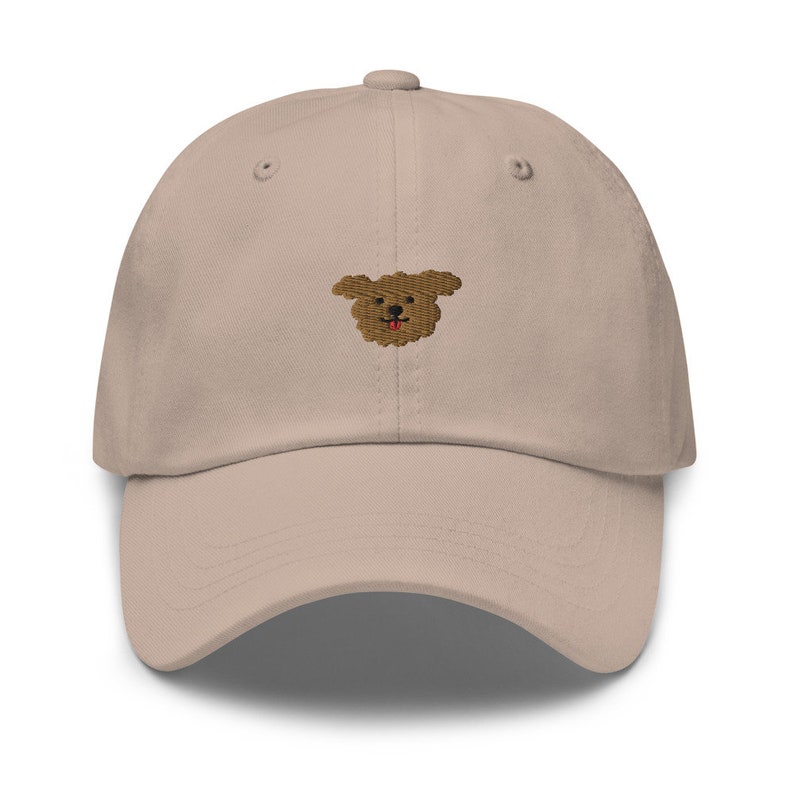 K&ouml;nnte beinhalten: Eine beige Baseballkappe mit einem gestickten braunen Hundegesicht auf der Vorderseite.