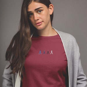 May include: A maroon t-shirt with a small embroidered image of four figures walking in a line. The figures are wearing different colored clothing, including blue, black, and white.