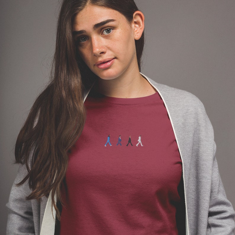 May include: A maroon t-shirt with a small embroidered image of four figures walking in a line. The figures are wearing different colored clothing, including blue, black, and white.