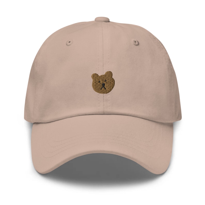May include: A pale pink baseball cap with a curved peak and adjustable strap. It has a small, embroidered brown bear face on the front. The hat is made of a soft material and has a classic, casual design.