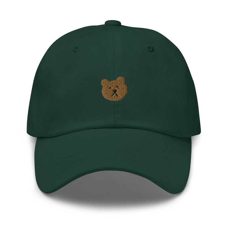 May include: A dark green baseball cap with a curved peak. The cap has a small, embroidered bear face in light brown on the front. The hat is made of a soft fabric and has a classic, adjustable design.