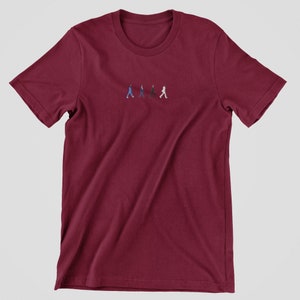 May include: A maroon t-shirt with a small embroidered image of four figures walking across a street, reminiscent of the iconic Abbey Road album cover.