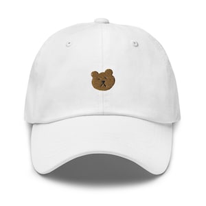 May include: White baseball cap with a curved peak. The cap has a small, embroidered brown bear face on the front. It has a classic six-panel design with a button on top and an adjustable strap at the back.