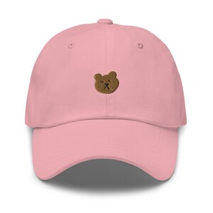 May include: A pale pink baseball cap featuring a brown embroidered bear face. The cap has a curved peak and an adjustable fabric strap at the back. The hat is made from a soft, durable material.