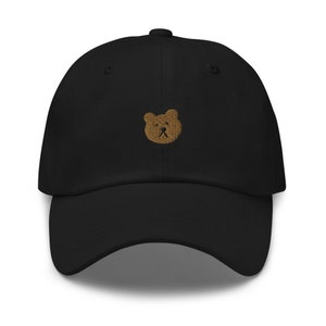May include: Black baseball cap with a curved peak and adjustable strap. The cap has a small, embroidered bear face in a light brown colour on the front. The hat is made of a soft, durable fabric.