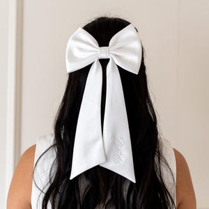 May include: A white satin bow with long tails, the tails are embroidered with the word "Bride".