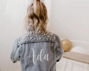 May include: A light blue denim jacket featuring a back panel embellished with numerous white pearls. The name Ada is printed in a white cursive script across the lower back section of the jacket. The garment has a classic collar and long sleeves.