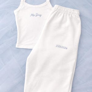 Puede incluir: Conjunto de pijama blanco con camiseta de tirantes y pantalón de chándal. La camiseta de tirantes tiene tirantes finos y el texto "Mrs. Diaz" en escritura azul claro. El pantalón de chándal tiene la fecha "2/28/2026" en azul claro.